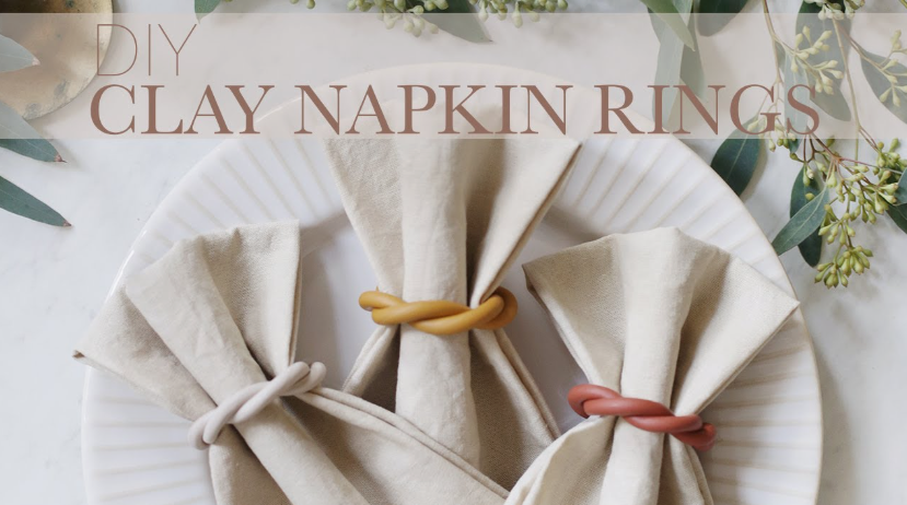 An overhead shot of three beige cloth napkins neatly folded on a white plate and secured with twisted air dry clay crafts styled as napkin rings in cream, mustard yellow, and terracotta colors.