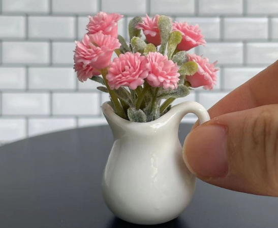 A close-up of delicate air dry clay crafts shaped into a miniature white pitcher holding a bouquet of tiny pink flowers, shown next to a hand against a white tile background.