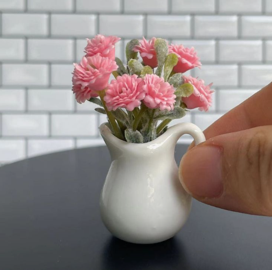 A close-up of delicate air dry clay crafts shaped into a miniature white pitcher holding a bouquet of tiny pink flowers, shown next to a hand against a white tile background.