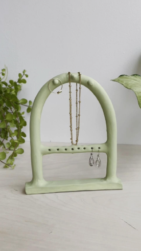 A pale green arch-shaped jewelry holder showcasing creative air dry clay ideas with small pegs holding a ring, a necklace, and earrings.