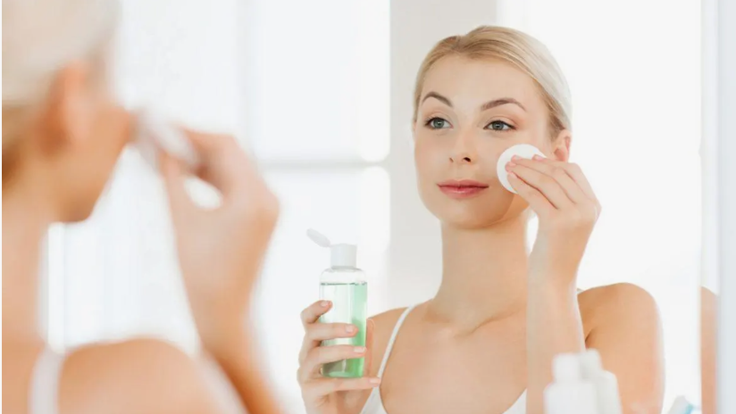 "A woman using a cotton pad and toner to cleanse her face in front of a mirror during her Sunday beauty reset routine."