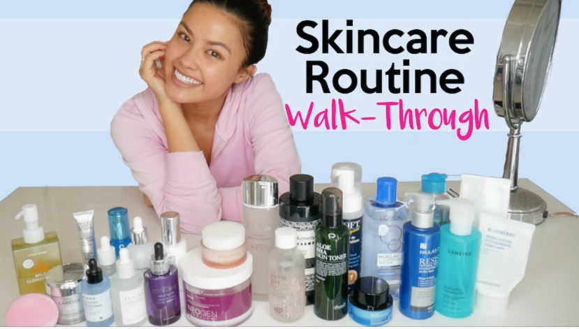"A smiling woman in a pink top surrounded by a wide assortment of skincare bottles and products during a Sunday beauty reset routine walkthrough."