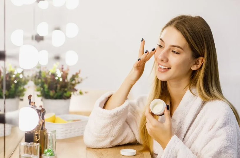 "A smiling woman in a plush white bathrobe applying facial cream from a small jar during her Sunday beauty reset routine in front of a lighted vanity mirror."