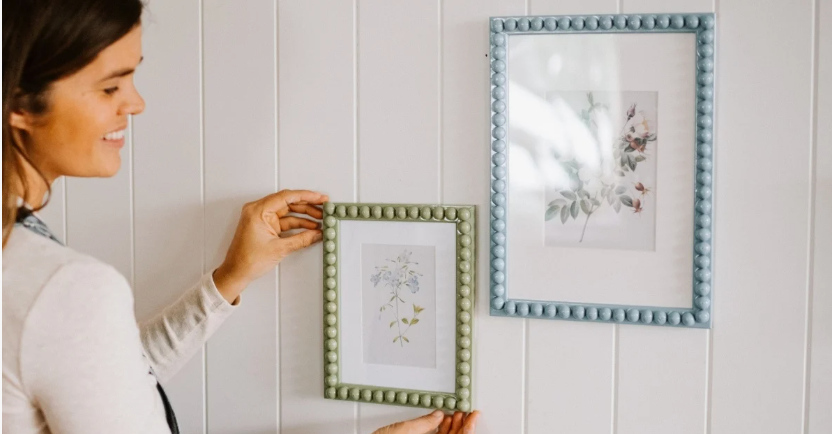 "A woman hanging beaded picture frames on a white paneled wall as part of 25 easy DIY home decor crafts for wall art."