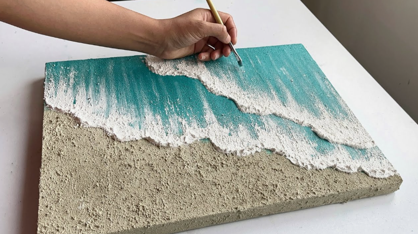 "A pair of hands using a brush and textured medium to create a 3D ocean wave canvas art, featured as one of 25 easy DIY home decor crafts."