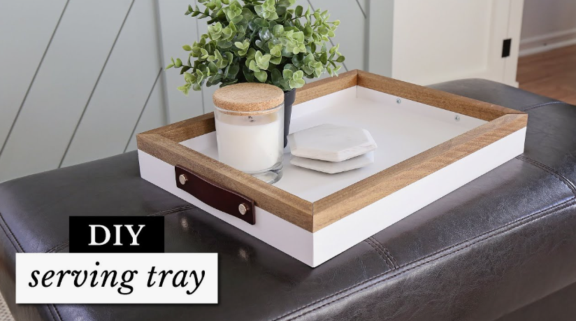 "A modern wood and white serving tray with leather handles holding a candle and coasters, featured as one of 25 easy DIY home decor crafts for ottoman styling."