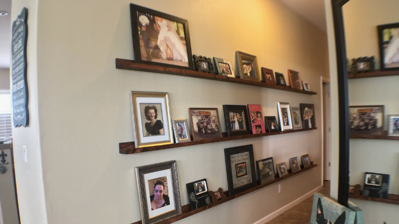 "A hallway with three long floating wooden shelves displaying a curated collection of framed family photographs, featured as part of 25 easy DIY home decor crafts for gallery wall styling."