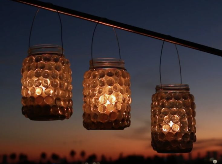 "Three hanging glass jar lanterns decorated with glass gems and glowing candles against a twilight sky, showcased as part of 25 easy DIY home decor crafts for outdoor ambiance."