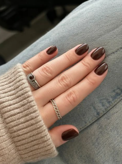 A hand with short, glossy dark brown nails wearing silver rings, styled for fall nail ideas 2026.
