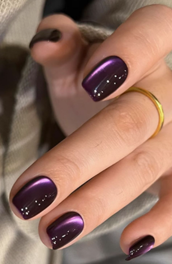 A hand with short, magnetic shimmer dark purple nails wearing a gold ring, styled for fall nail ideas 2026.