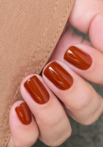A hand with short, glossy burnt orange and terracotta nails next to a camel-colored fabric, styled for fall nail ideas 2026.