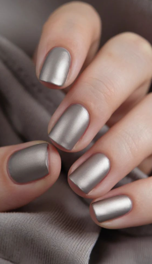 A hand with short, metallic satin-finish grey-taupe nails against a soft fabric background, styled for fall nail ideas 2026.