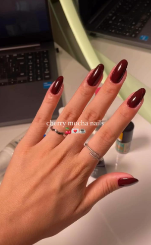 A hand with long, glossy cherry mocha nails and silver rings over a desk setting, styled for fall nail ideas 2026.
