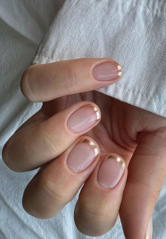 A hand with short, squoval nails featuring metallic gold French tips against a white fabric background, styled for fall nail ideas 2026.