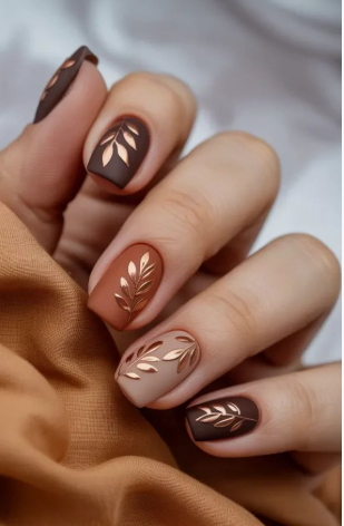 A hand with short, matte autumn-toned nails featuring metallic gold leaf branch art, styled for fall nail ideas 2026.