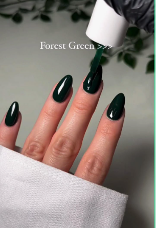 A hand with long, glossy forest green almond nails having polish applied, styled for fall nail ideas 2026.