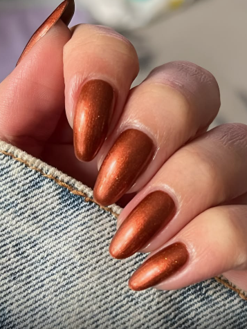 A hand with long, metallic bronze and copper almond-shaped nails against denim, styled for fall nail ideas 2026.