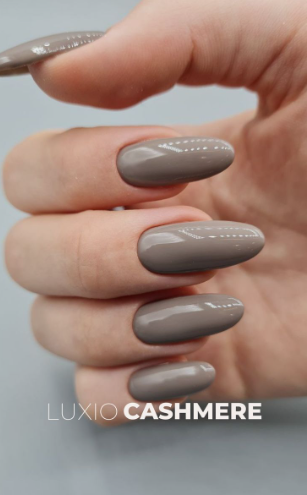 A hand with long, glossy taupe "Luxio Cashmere" almond nails, styled for fall nail ideas 2026.