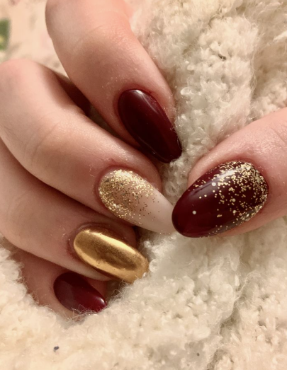 A hand resting against a cozy white sweater displaying almond-shaped nails with deep burgundy and sparkling gold glitter accents, styled for fall nail ideas 2026.