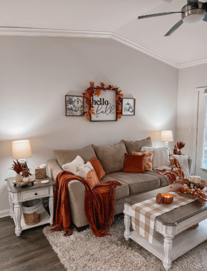 A cozy living room couch styled with warm rust-colored blankets, throw pillows, and a "Hello Fall" wall sign, highlighting creative fall decor ideas without pumpkins.
