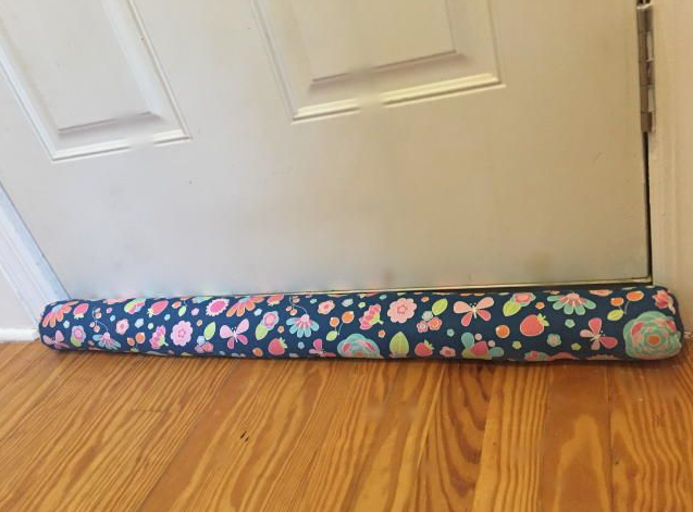 20 DIY Crafts With Old Clothes That Are Easy to Make A floral fabric draft stopper placed at the bottom of a door on a wooden floor, showcasing clever DIY crafts with old clothes for home insulation.