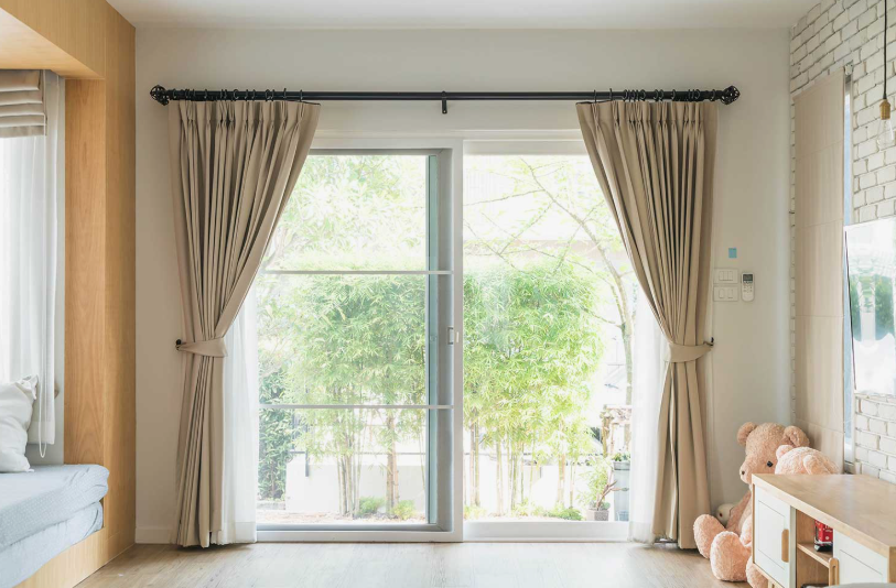 "Cozy home decor ideas for a small apartment featuring a bright sliding glass door with layered beige curtains, a black curtain rod, and a cute teddy bear sitting beside a wooden media console."