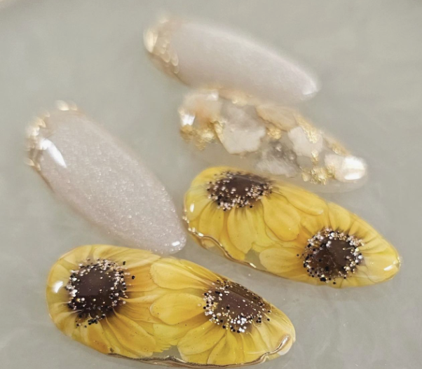 24 Sunflower Nail Ideas for a Bright, Cheerful Manicure sunflower nail ideas featuring press-on nails with encapsulated yellow sunflowers and dark centers accented with glitter, paired with neutral white shimmer and textured crystal stone embellishments.