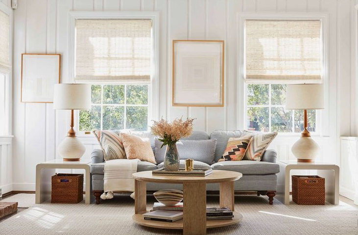"Cozy home decor ideas for a small apartment featuring a bright living room with a grey sofa, matching side lamps, wooden coffee and end tables, and framed art against white paneled walls."