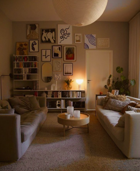 "Cozy home decor ideas for a small apartment featuring a warm, eclectic living room with two facing couches, a round coffee table, low bookshelves, a gallery wall, and soft ambient lighting."