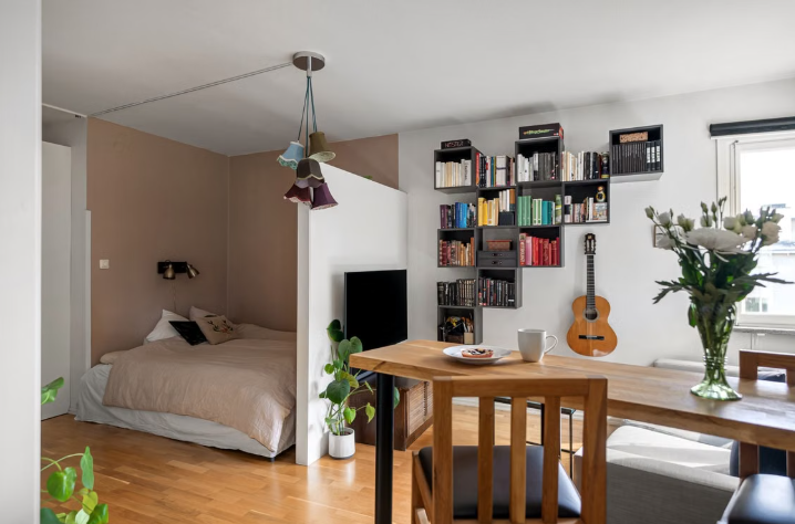 "Cozy home decor ideas for a small apartment featuring a functional studio layout with an open bedroom alcove, a wooden dining table, and a geometric wall-mounted bookshelf display."