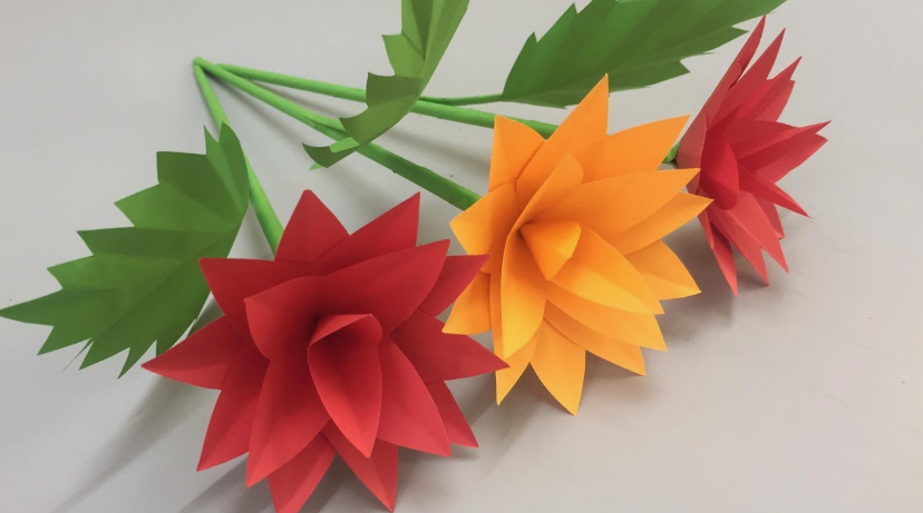 Three handmade paper flowers on green stems with jagged green leaves, featuring layered, pointed petals in red, orange, and another red flower in the background.