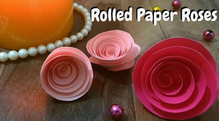 Three handmade paper roses in varying shades of pink and light peach, featuring spiral rolled petals arranged on a wooden surface alongside decorative pearls and text reading "Rolled Paper Roses".