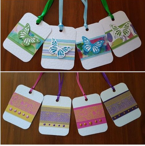 Two rows of handmade white paper gift tags featuring decorative ribbon loops. The top row displays tags adorned with colored butterfly cutouts over patterned background stripes, while the bottom row features tags decorated with patterned washi tape and small accent beads.