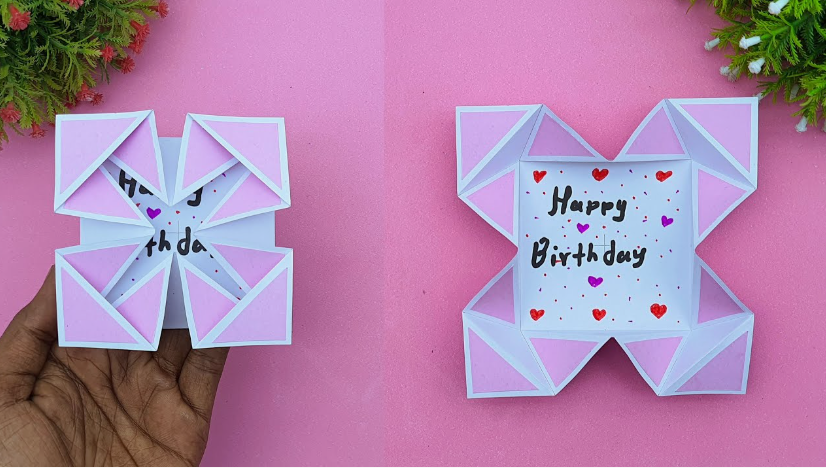 A handmade origami folding card made of white paper with pink triangular fold details, shown being held closed on the left and unfolded on the right to reveal the handwritten message "Happy Birthday" decorated with small hearts.