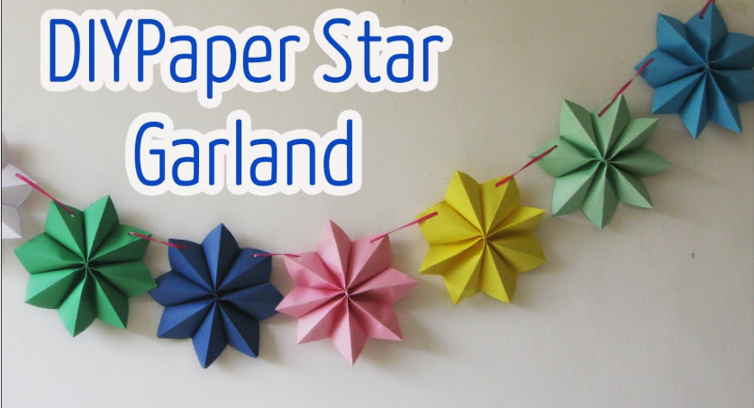 A colorful hanging garland made of pleated accordion paper stars in shades of green, blue, pink, yellow, and light blue, strung together on a thin red cord against a white wall, accompanied by text reading "DIY Paper Star Garland".