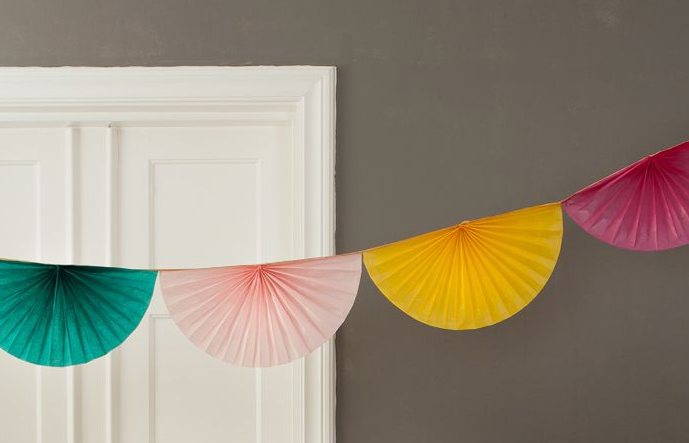 Two draped strings of colorful, semicircular pleated paper fan decorations hanging above a white bed frame with patterned and solid-colored pillows.