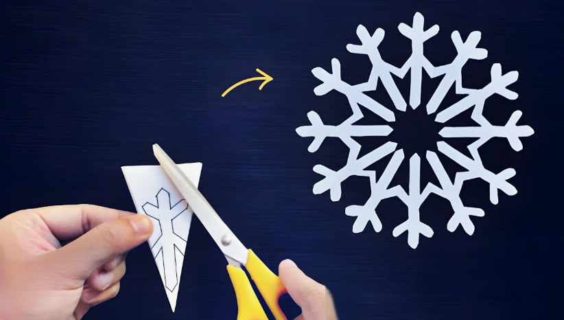 A pair of yellow-handled scissors cutting along an outlined template on a folded white triangular paper piece, with an arrow pointing to the fully unfolded, intricate white paper snowflake design against a dark surface.
