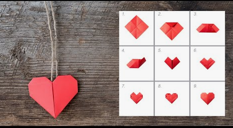 A step-by-step visual guide numbered one through nine showing the folding sequence to create an origami heart from a square piece of red paper, accompanied on the left by a finished red paper heart hanging from a rustic twine string against a wooden background.   