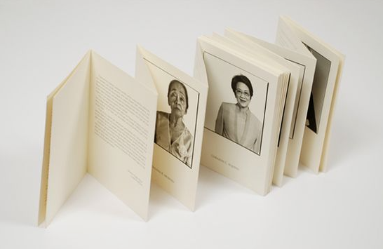  A handmade accordion-fold paper booklet standing upright on a white surface, featuring cream-colored pages with printed text blocks and framed black-and-white portrait photographs.   
