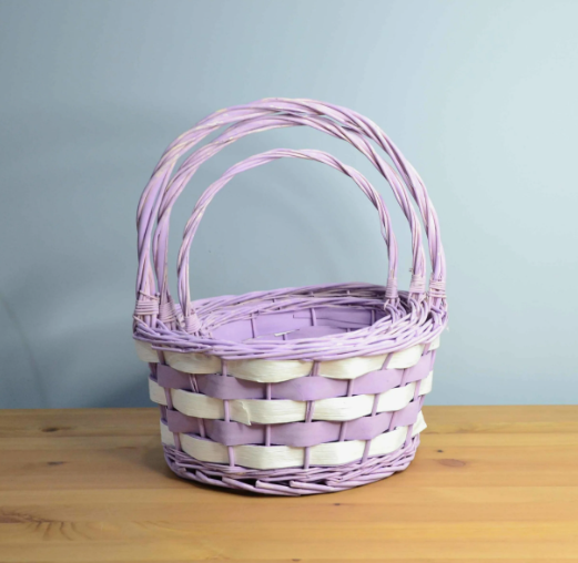 A pair of nested handmade woven baskets crafted from light purple and white paper strips, featuring rounded bases and arched braided handles, sitting on a wooden surface against a pale blue background.