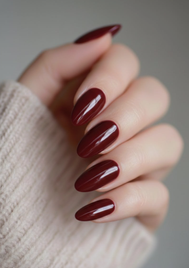 Glossy oval-shaped burgundy nail designs featuring a rich, solid dark red manicure paired with a cozy beige sweater.