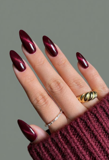 Stunning almond-shaped burgundy nail designs featuring a mesmerizing cat-eye gel effect with deep dark red shimmer and gold rings.
