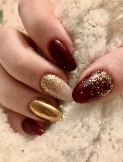 Festive burgundy nail designs featuring a mix of solid dark red polish, a metallic gold accent nail, and glitter-gradient details holding a soft white knit fabric.