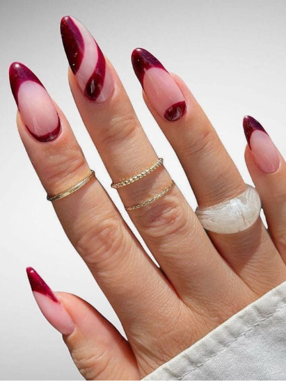 Artistic almond-shaped burgundy nail designs showcasing unique diagonal swirl patterns and deep dark red French tips on a nude base.