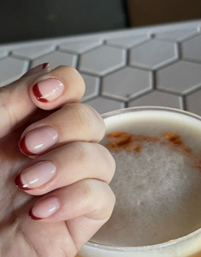 Classic short oval burgundy nail designs featuring clean, delicate dark red French tips on a natural nude base next to a coffee cup.   