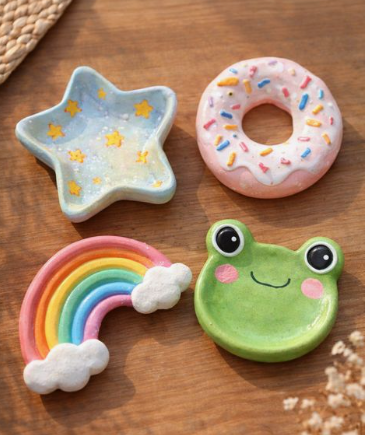 An overhead shot of four colorful air dry clay crafts arranged on a wooden table, including a blue star dish, a pink frosted donut, a rainbow with white clouds, and a green smiling frog dish.