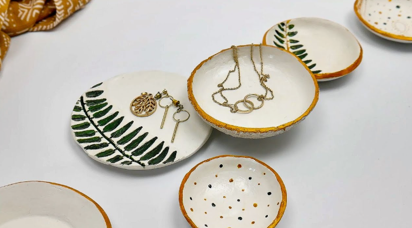 A flatlay of several handmade air dry clay crafts featuring white trinket dishes decorated with green fern prints, gold rims, and polka dots, some holding delicate jewelry pieces.