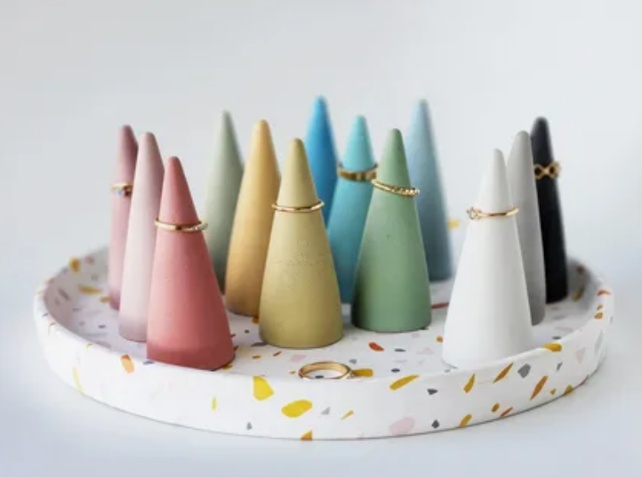 A collection of colorful air dry clay crafts styled as cone-shaped ring holders, organized on a matching white terrazzo-patterned tray with several rings placed on them.