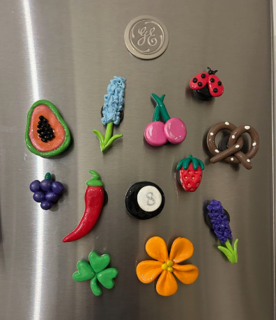 A collection of colorful air dry clay crafts shaped into miniature magnets displayed on a refrigerator, featuring foods, flowers, a ladybug, and an eight-ball.