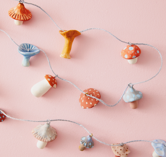 A close-up of colorful air dry clay crafts shaped into miniature mushrooms strung together on strings to form decorative garlands against a pink background.